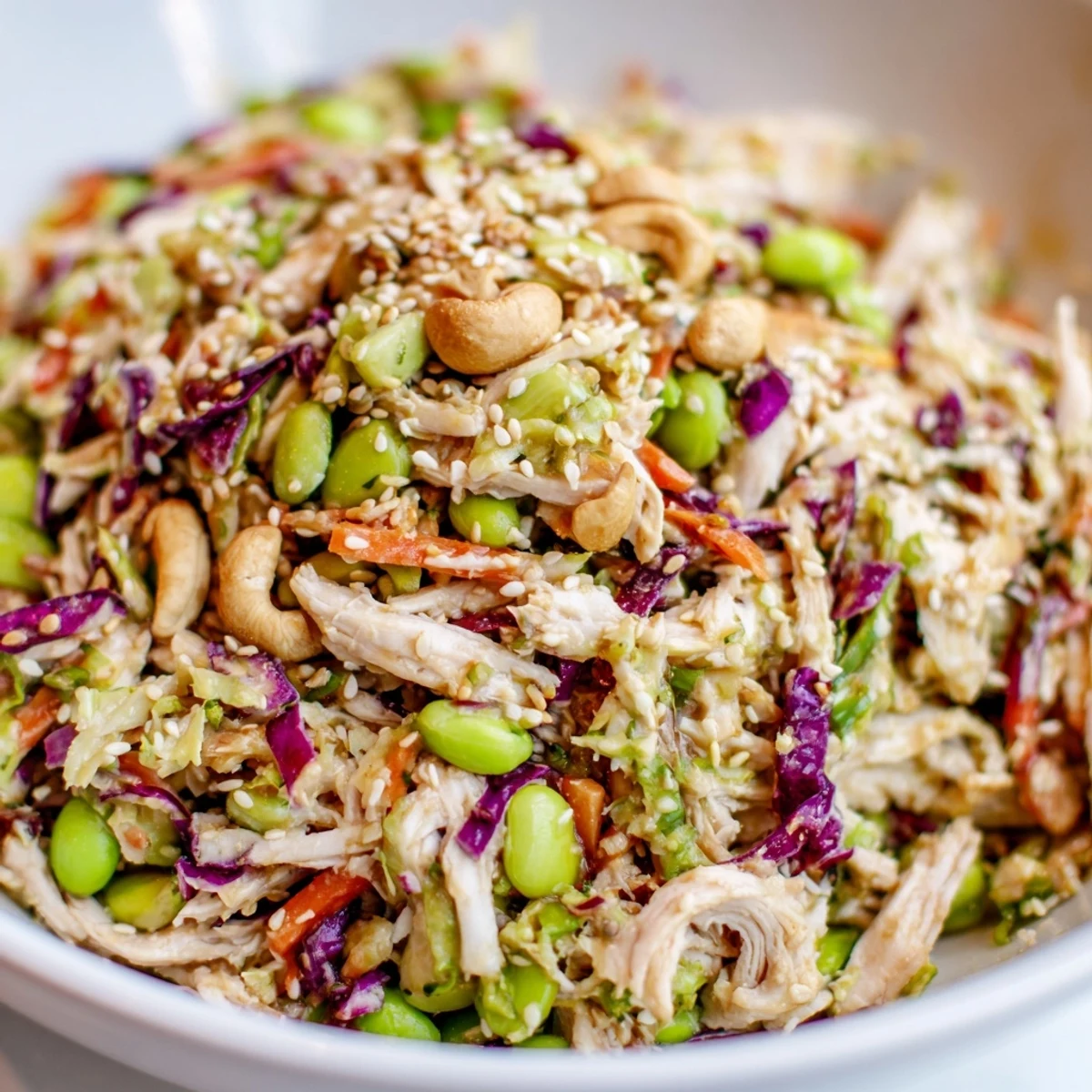 A vibrant bowl of Edamame Crunch Chicken Salad tossed with crisp cabbages, carrots, and roasted cashews on a wooden table.  