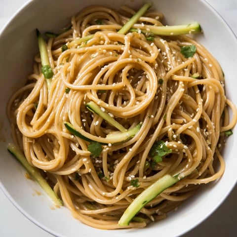 A close-up of a vibrant Spicy Sesame Noodle Salad, showcasing glossy noodles coated in a reddish chili oil dressing, topped with julienned cucumber, fresh cilantro, and toasted sesame seeds.  